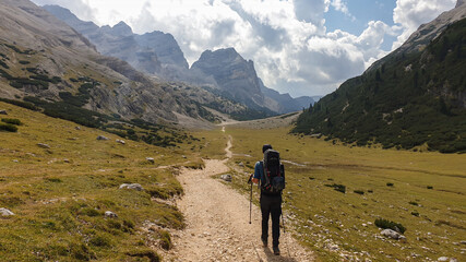 A man with a hiking backpack hiking on a gravelled road in high Italian Dolomites. There is a dense forest at the foothill, and steep and sharp mountain chain in the back. Discovering and exploring