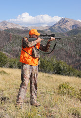 adult deer hunter wearing orange and aiming scoped rifle in mountains