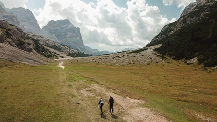 Fototapeta na wymiar A couple hiking along a pathway leading through a valley in Italian Dolomites. High, sharp mountains around. Stony and raw landscape, green meadow. Remote and desolate place. Freedom of exploration
