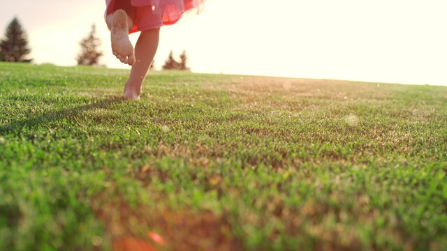 Unknown Girl Playing In City Park. Rear View Of Kid Legs Running On Meadow.