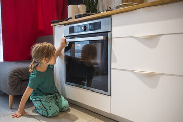 Fototapeta na wymiar Curious child watching through glass of kitchen oven in a home with white interior. Adorable toddler girl playing in the kitchen next to a modern white oven helping by cooking and baking an apple pie