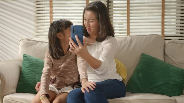 Happy Asian Family mum and little kid daughter using a smart phone and having a video call chatting or taking a selfie sitting on a couch fun and enjoying online communication in living room at home.