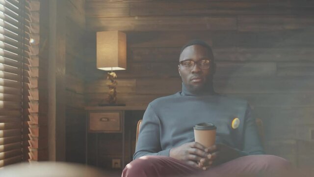Portrait of young African American man in smart casual outfit and glasses sitting on chair, holding disposable coffee cup and posing for camera with confidence