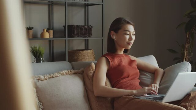 Young Asian woman in wireless earphones sitting on sofa in living room, waving and talking via online video call on laptop