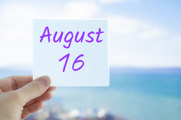 August 16th. Hand holding sticker with text August 15 on the blurred background of the sea and sky. Copy space for text. Month in calendar concept