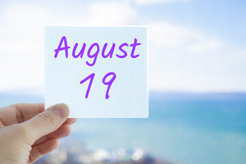 August 19th. Hand holding sticker with text August 15 on the blurred background of the sea and sky. Copy space for text. Month in calendar concept