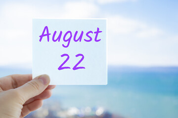 August 22nd. Hand holding sticker with text August 15 on the blurred background of the sea and sky. Copy space for text. Month in calendar concept
