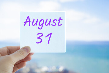 August 31st. Hand holding sticker with text August 15 on the blurred background of the sea and sky. Copy space for text. Month in calendar concept