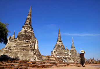 Fototapeta premium Female Visitor in White Hat Walking along the Incredible Historic Pagoda Ruins of Wat Phra Si Sanphet in Ayutthaya, Thailand