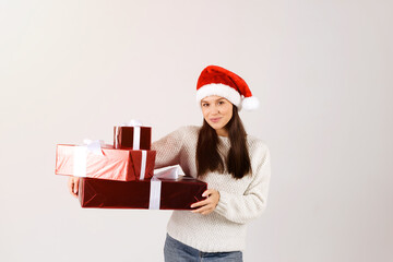 Woman in white sweater holding many gift christmas boxes wearing red santa hat isolated over gray background
