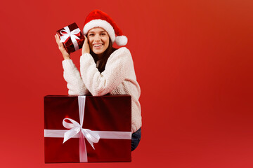 Happy pretty young woman in red santa hat holding a smal gift christmas box sitting near big red present box isolated over red background