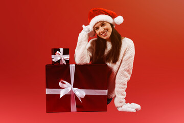 Happy pretty young woman in red santa hat sitting near big red gift box isolated over red background wearing white knitted mittens