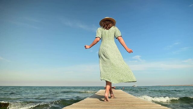A young woman in a green summer dress and a straw hat in slow motion walks along a wooden pier in the morning on the sea during the wind. Walk grace beauty and femininity
