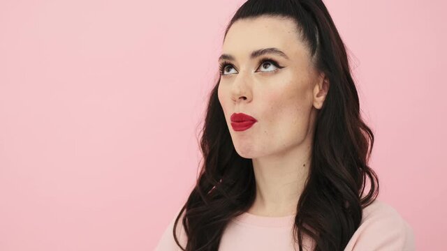 An attractive woman with red lips is eating bubblegum standing isolated over a pink background in the studio