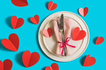 Served table with plate and cutlery forcelebration of Valentine's Day. On plate is napkin with paper heart. Flatlay on bright blue background with red hearts. Top view.