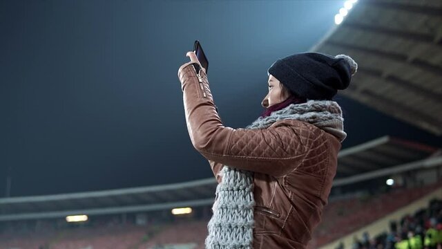Girl dressed in hat and scarf shooting video at the stadium. Gadget in the hands of young woman. Shooting interesting moments in the video with the help of technology. Concept of modern technologies