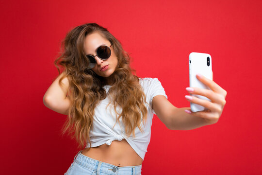 Closeup of sexy amazing beautiful young woman holding mobile phone taking selfie photo using smartphone camera wearing round sunglasses everyday stylish outfit isolated over colorful wall background