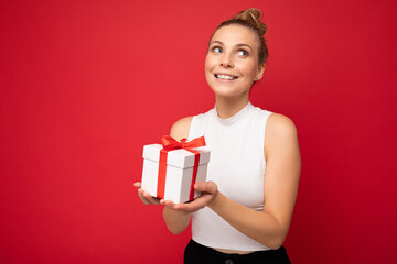 Charming positive smiling young blonde woman isolated over red background wall wearing white top holding gift box and looking up and dreaming