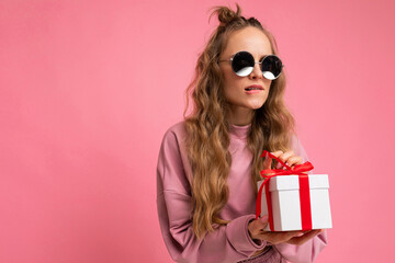 Shot of beautiful positive young blonde curly woman isolated over pink background wall wearing pink sport clothes and sunglasses holding gift box and unboxing surprise looking to the side