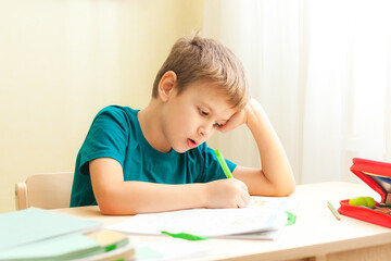 7 yeas old boy sitting at desk and doing homework. Child write notes in notebook, difficult homework, child concentrating on examples