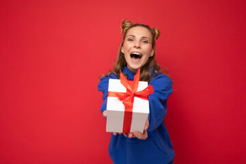 Emotional attractive happy young blonde woman isolated over red background wall wearing blue trendy hoodie holding gift box and looking at camera and shouting