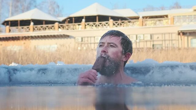 Cryotherapy. Young caucasian man with icy beard calmly eats an ice cream being emerged in the cold icy water