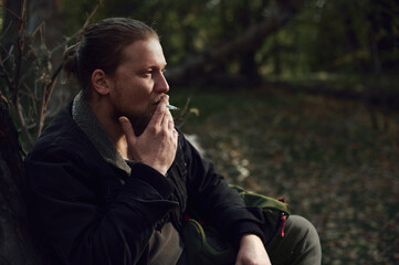 Side portrait of a smoking young bearded man adventurer relaxing in the autumn forest