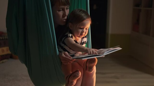 Baby and mother read a hammock. Mother reads a book with together girl being in the hammock at home