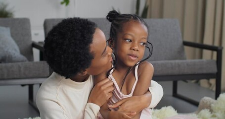 Beautiful woman in short hair black lady mom kisses little daughter afro american funny girl beloved child on cheek tenderly lovingly hugs child enjoys motherhood parenting at home sitting on floor