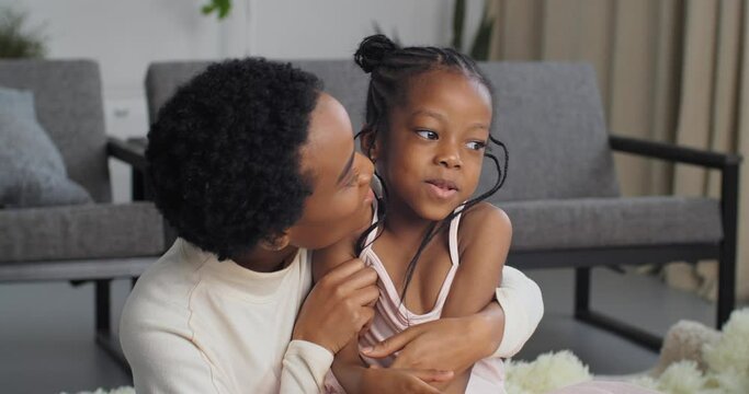 Beautiful woman in short hair black lady mom kisses little daughter afro american funny girl beloved child on cheek tenderly lovingly hugs child enjoys motherhood parenting at home sitting on floor