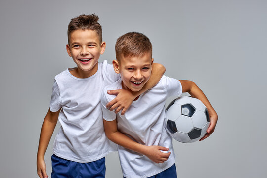 kid boys go in for soccer sport professionally, stand hugging each other, have fun, smile at camera, holding ball in hands, isolated portrait