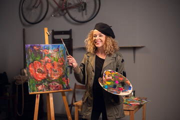 Fototapeta premium Young happy smiling female artist in an art workshop. Draws a beautiful abstract picture with flowers