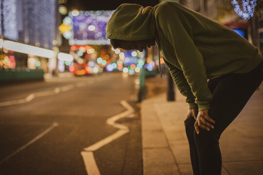 Young female runner resting with hands on knees on city street at night