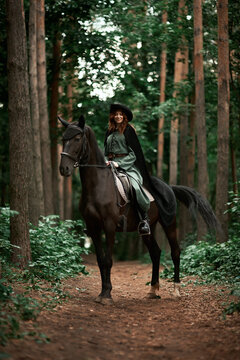 Young beautiful woman in a suit, green dress, corset, black topm hat on a black horse on nature in the forest. fairy tale, creative photo session of a girl with a horse