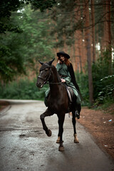 Fototapeta premium Young beautiful woman in a suit, green dress, corset, black topm hat on a black horse on nature in the forest. fairy tale, creative photo session of a girl with a horse