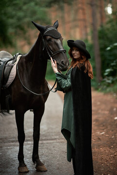 Young beautiful woman in a suit, green dress, corset, black topm hat on a black horse on nature in the forest. fairy tale, creative photo session of a girl with a horse