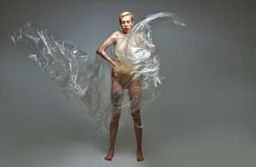 portrait of slender caucasian female in bodysuit posing in polyethylene bag, woman in gray studio background. ecological problem, zero waste