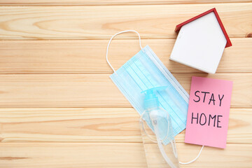 Inscription Stay Home on pink paper with medicine mask, sanitizer bottle and wooden house on brown backgorund