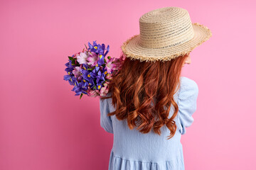 redhead female in summer hat and dress holding flowers, posing, isolated on pink background