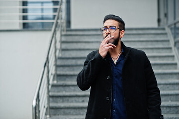 Middle eastern entrepreneur wear black coat and blue shirt, eyeglasses against office building smoking cigarette.