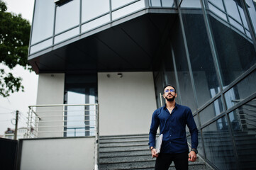 Middle eastern entrepreneur wear blue shirt, eyeglasses with laptop against office building.