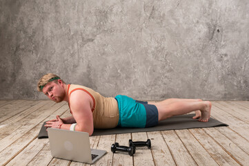 Funny fat man in sportswear doing sports exercises looking into a laptop. home workout 