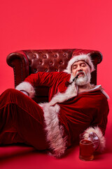 santa man in suit lie on the floor near chair with smoking pipe and beer isolated over red background