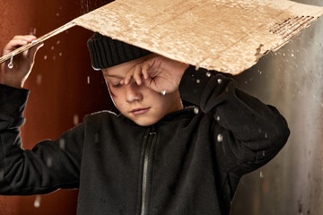 homeless child use cardboard so as not to get wet, while raining outdoors, he is crying