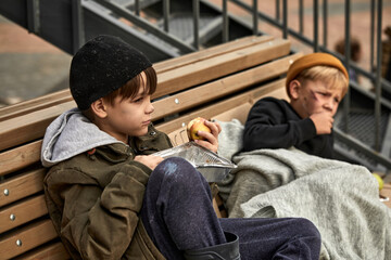 Fototapeta premium two homeless boys sit eating food on the bench, little caucasian children, street kids are hungry, get some food by kind people