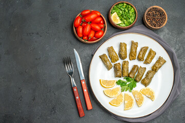 top view stuffed grape leaves on white oval plate bowls with cherry tomatoes parsley black pepper knife and fork and purple kitchen towel on dark background with copy space