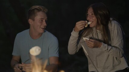 Rack focus of teenage couple roasting and eating marshmallows over campfire at night / Tibble Fork, Utah, United States - Powered by Adobe
