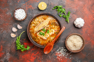 top view homemade chicken and rice soup in a bowl wooden spoon bowls with sea salt and rice garlic parsley leaves slice of lemon on dark red background