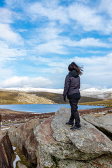 Obraz premium Vertical view of a white Caucasian woman on a rock overlooking a landscape of a lake and the snowy mountains in the background on a sunny day
