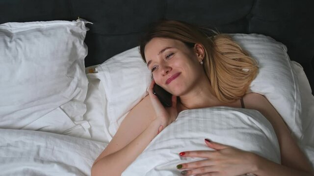 Top view of happy caucasian woman talking on mobile phone while lying in bed at home.